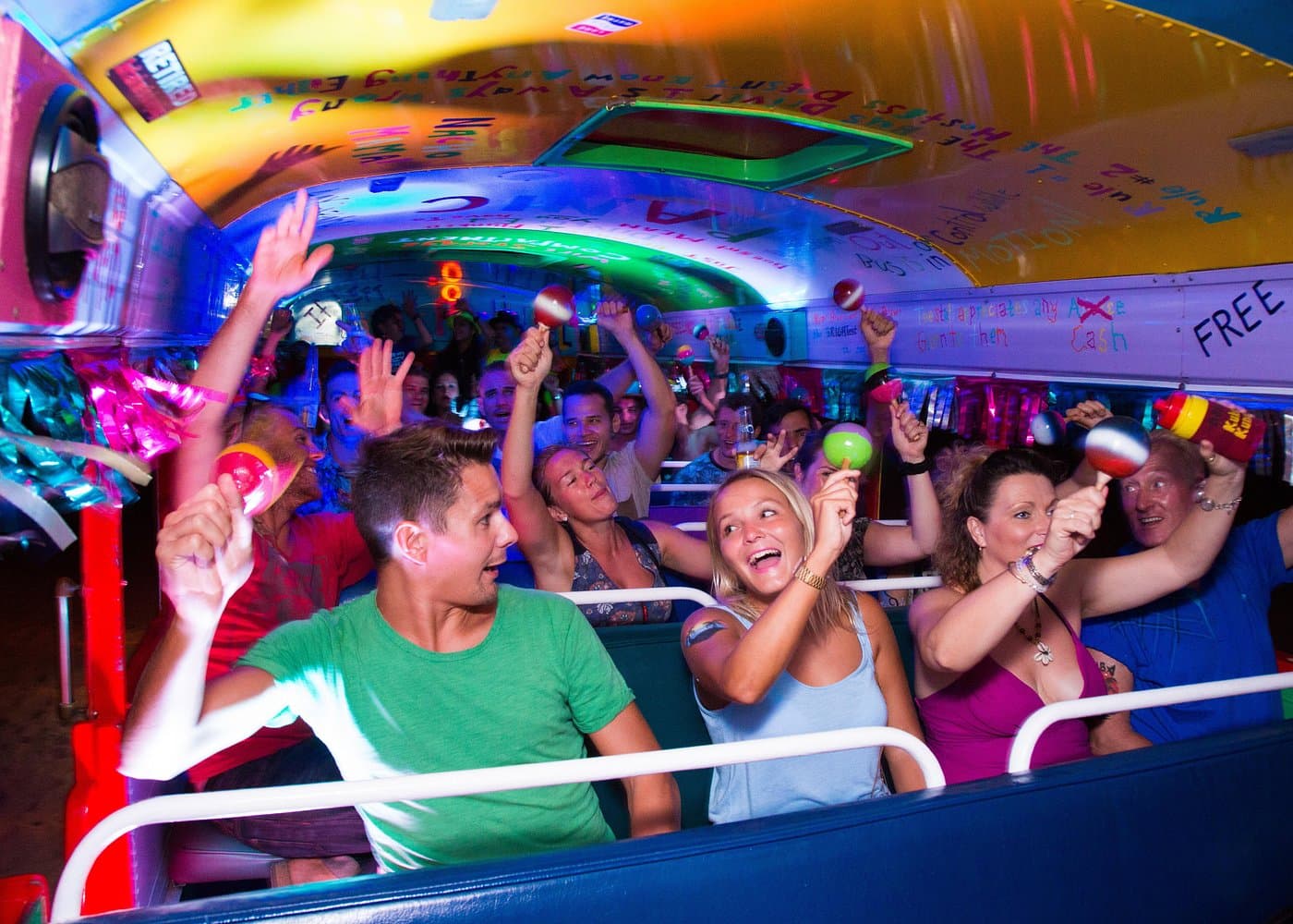 Guests laughing on a Kukoo Kunuku party bus at night, shaking maracas under colored lights.